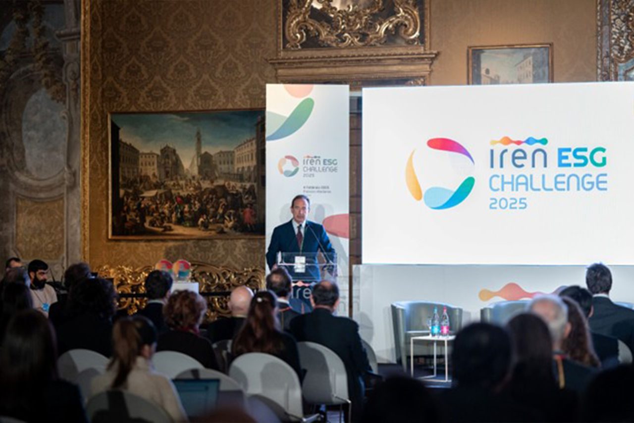 in abito scuro parla da un podio trasparente durante l'evento "Iren ESG Challenge 2025" a Palazzo Madama, Torino. Sullo sfondo, un grande schermo e un banner mostrano il logo colorato dell'evento. La sala è decorata con eleganti dettagli dorati e dipinti storici, mentre il pubblico ascolta attentamente.