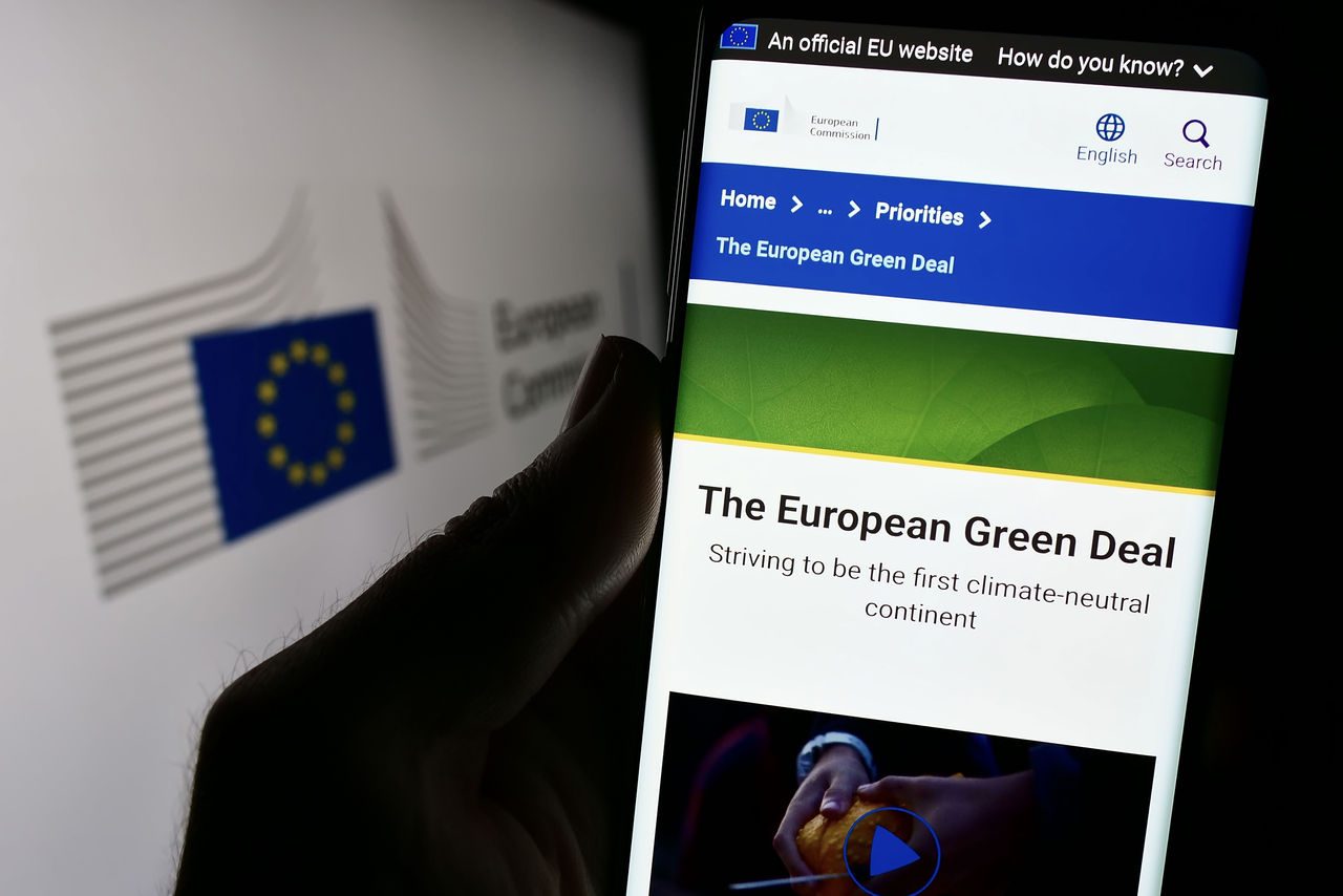 Schermo di smartphone che mostra il sito ufficiale della Commissione Europea dedicato al Green Deal, con il testo "The European Green Deal" e lo slogan per la neutralità climatica. Sullo sfondo, il logo della Commissione Europea sfocato.