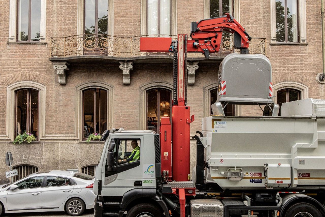 Camion per la raccolta dei rifiuti in città mentre solleva un cassonetto con un braccio meccanico. L’autista, con giubbotto catarifrangente, è visibile nella cabina di guida. Sullo sfondo, un edificio storico con finestre decorate e balconi in ferro battuto.