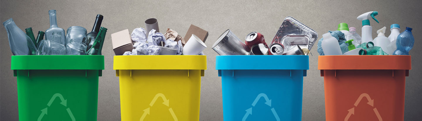 Quattro bidoni per la raccolta differenziata in fila, ciascuno con un colore diverso: verde per il vetro, giallo per la carta, blu per i metalli e marrone per la plastica. Ogni contenitore è riempito con rifiuti corrispondenti alla sua categoria.