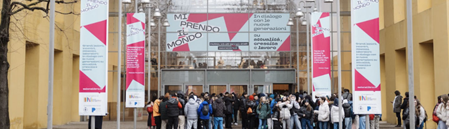 Ingresso di un edificio con un grande striscione che riporta il titolo “Mi prendo il mondo” e slogan dedicati al dialogo con le nuove generazioni su attualità, crescita e lavoro. Un folto gruppo di giovani in attesa all’ingresso, sotto banner verticali con la stessa grafica dell’evento.