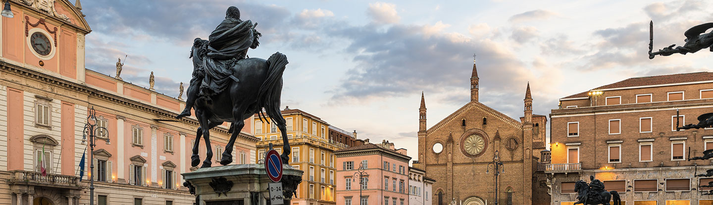 Panorama della città di Piacenza al tramonto con la statua equestre di Alessandro Farnese in primo piano e la Chiesa di San Francesco sullo sfondo, circondata da edifici storici illuminati.