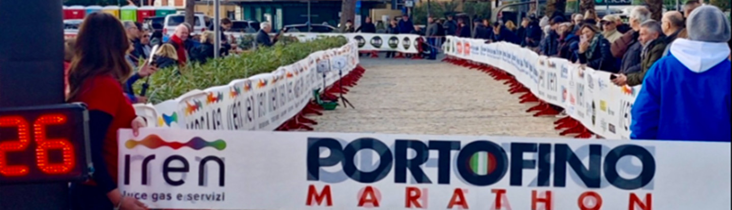 Arrivo della Portofino Marathon con striscioni di Iren lungo il percorso e spettatori allineati ai lati in una cornice mediterranea con palme.