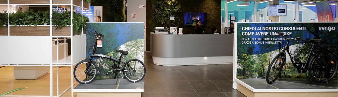 Spazio commerciale con espositori dedicati alla mobilità sostenibile, tra cui biciclette elettriche, e un'area verde interna con piante e pareti vegetali.