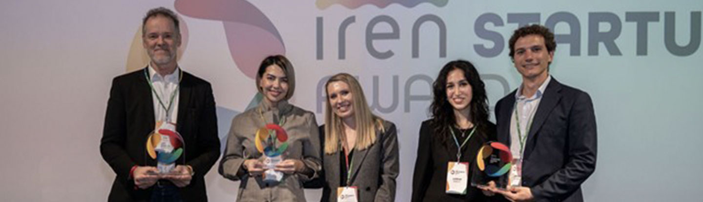 Le due vincitrici e una menzione speciale degli Iren Startup Awards 2025, sorridenti mentre tengono in mano i loro trofei. Sullo sfondo, il logo dell'evento proiettato su uno schermo.