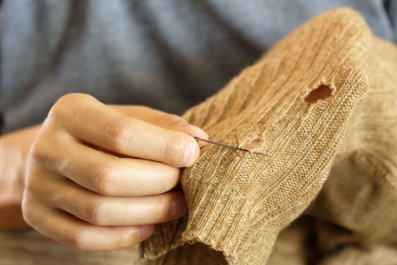 Primo piano di una mano che cuce a mano un maglione di lana marrone con un piccolo foro, simbolo di riparazione e riutilizzo nella moda sostenibile.