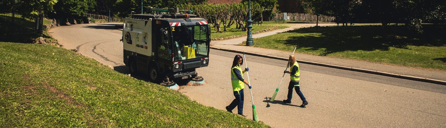 Due operatori ecologici con giubbotti catarifrangenti puliscono una strada in un’area verde, utilizzando scope, mentre una spazzatrice meccanica lavora nelle vicinanze.