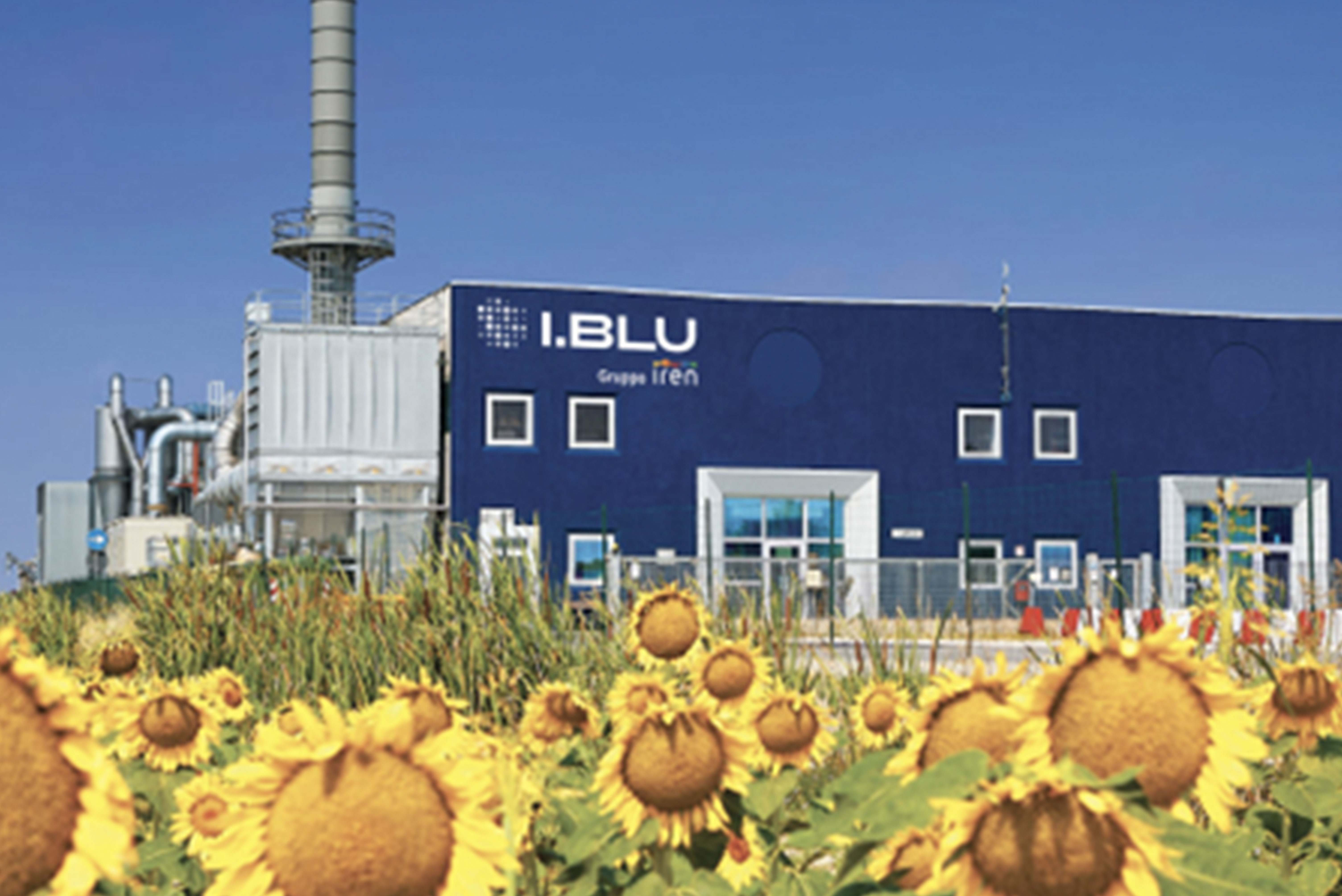 Stabilimento industriale del Gruppo Iren con la scritta "I.BLU" sulla facciata blu dell'edificio, circondato da un campo di girasoli sotto un cielo sereno.