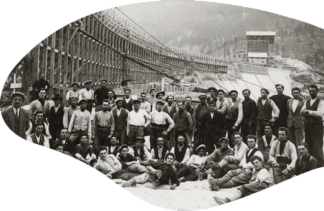 Foto storica in bianco e nero. Gruppo di operai in posa davanti alla diga di Ceresole Reale in costruzione.