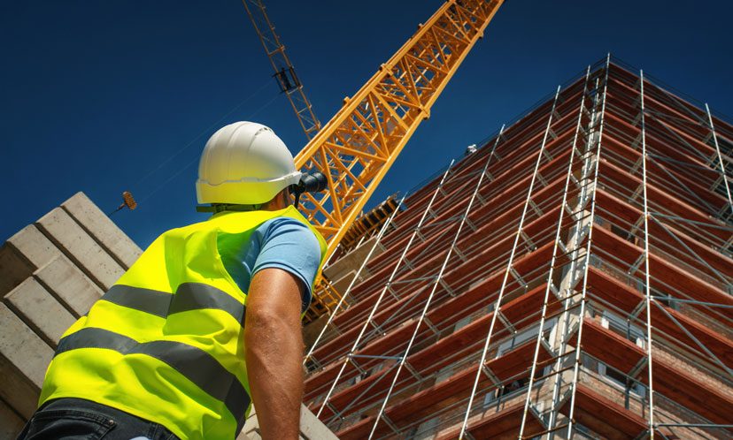 Vista dal basso di un dipendente tecnico con i dispositivi di protezione che guarda un cantiere edilizio con davanti una gru